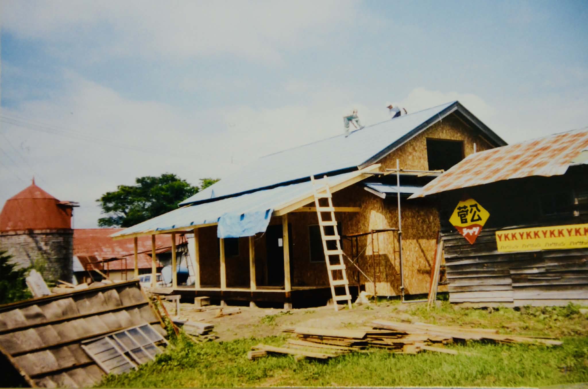 シゲチャンランドの建築風景。木造の建物の青い屋根の上では、2人の人物が作業をしており、手前にはたくさんの木材などの資材が置かれている